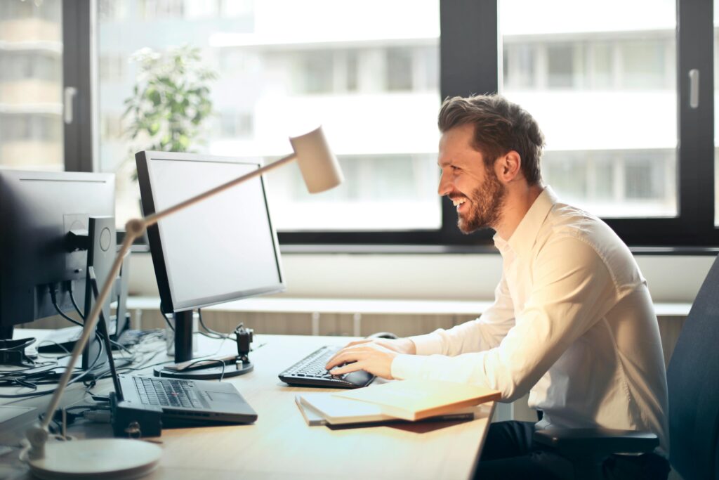 pexels-photo-840996-840996 A man smiling while working at an office desk with a computer and natural daylight streaming in through large windows.